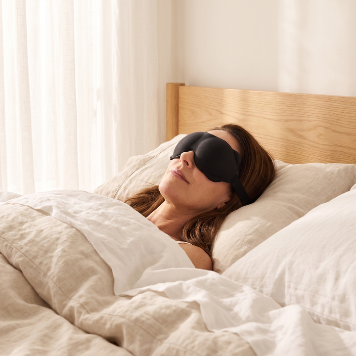 3D Contoured Sleep Mask in Black – worn by a woman lying in bed near sheer curtains