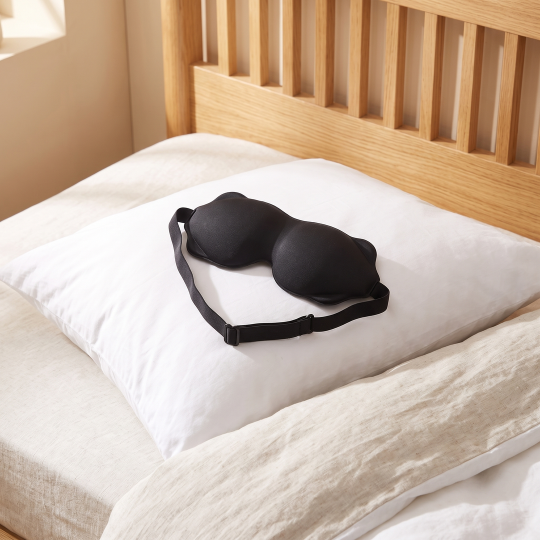 3D Contoured Sleep Mask in Black – resting on a white pillow on a bed with linen bedding
