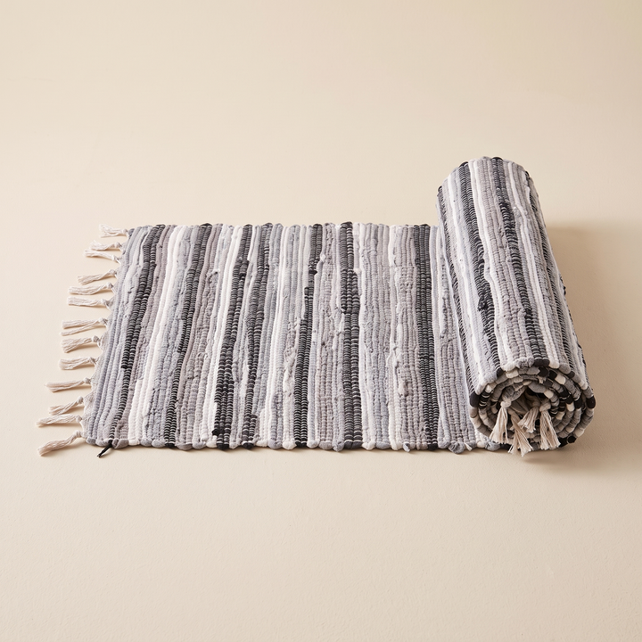 Boho Rag Rug – Woven Cotton Stripes in Gray – partially rolled up on a plain cream background, fringe visible