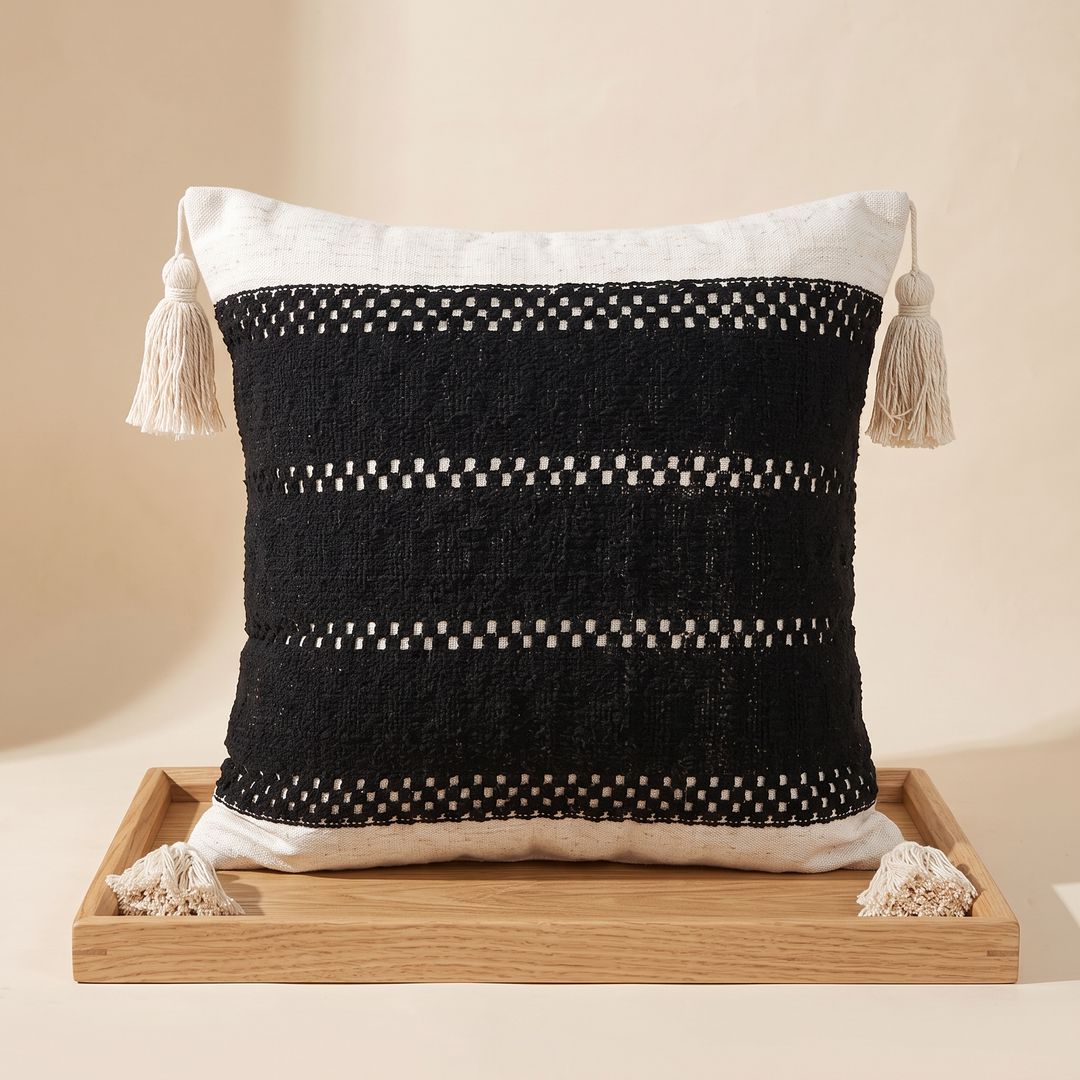 Boho Pillow Cover with Tassels – Woven in Black – Openwork – propped upright on a light wooden tray with tassels resting on its edge