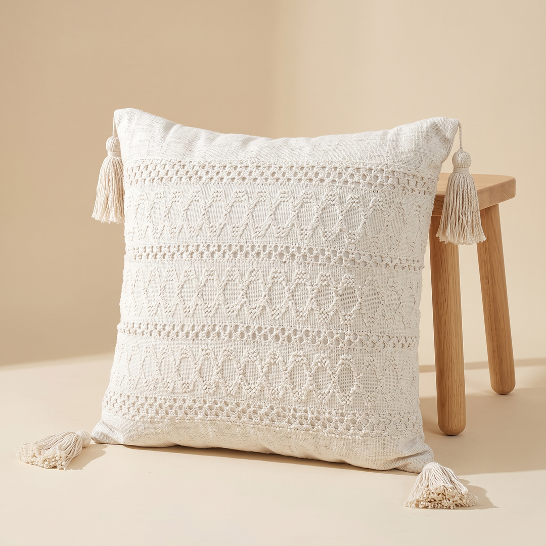 Boho Pillow Cover with Tassels – Woven in Cream – Openwork – leaning beside a wooden stool against a beige wall