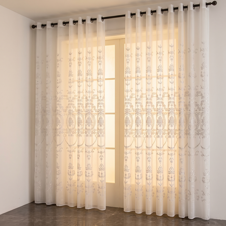 Embroidered Sheer Curtain Panel – Grommet Top in White – two panels hanging on a black rod beside a glowing window