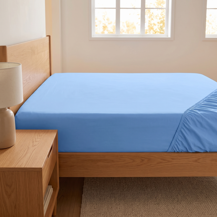 Fitted Sheet in Light Blue – side view on a bed beside a wooden nightstand with lamp