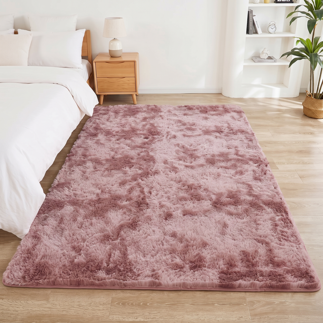 Fluffy Shag Rug in Dusty Pink – Tie-Dye – lengthwise along a bedside, lamp-topped nightstand and white shelves beyond