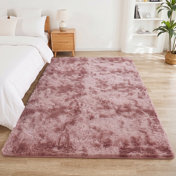Fluffy Shag Rug in Dusty Pink – Tie-Dye – lengthwise along a bedside, lamp-topped nightstand and white shelves beyond