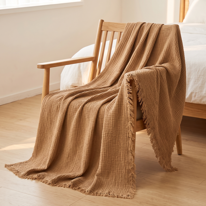 Crinkled Cotton Blanket with Fringe in Camel – draped over a wooden armchair beside a bed on a hardwood floor