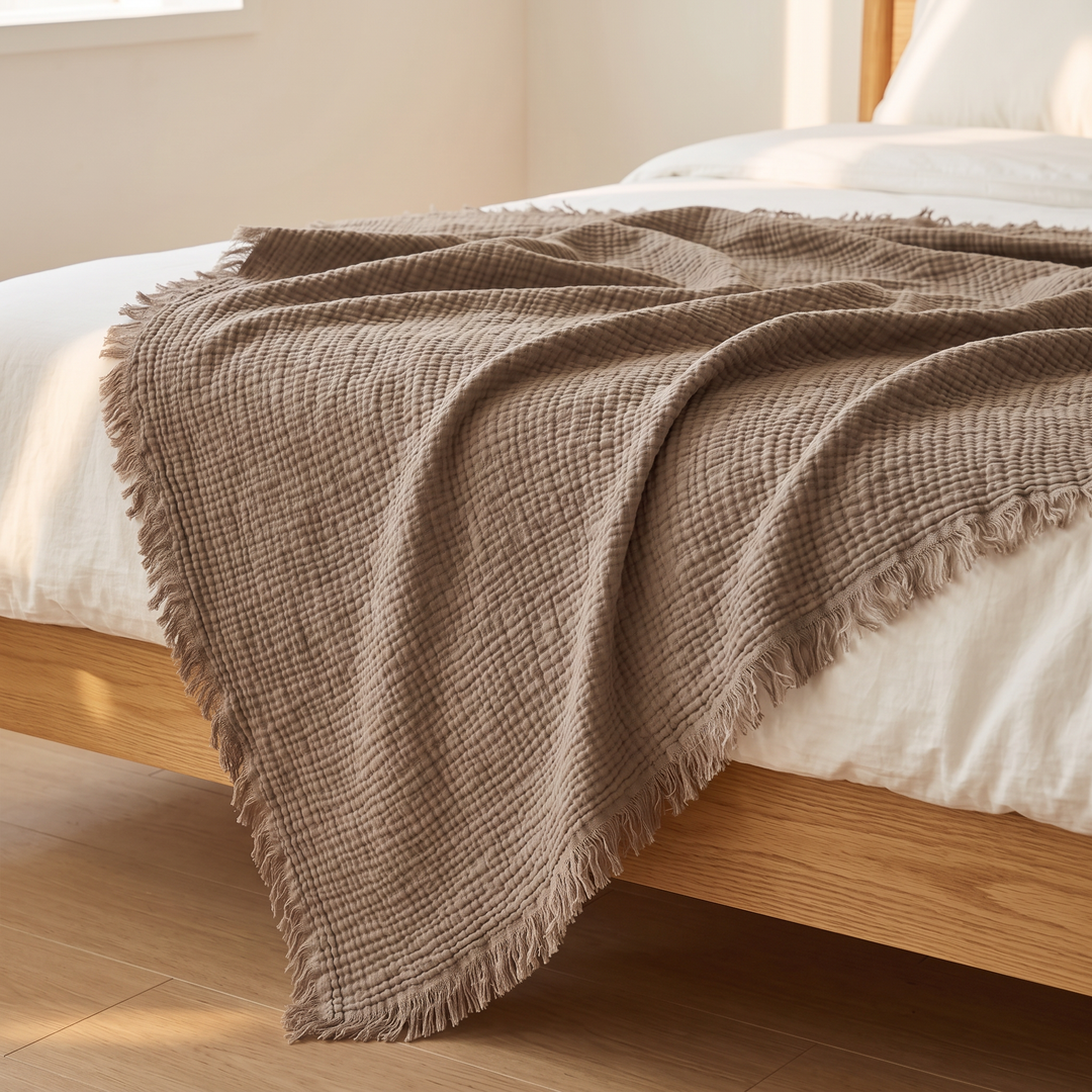 Crinkled Cotton Blanket with Fringe in Taupe – hanging over a wooden bed frame in sunlight