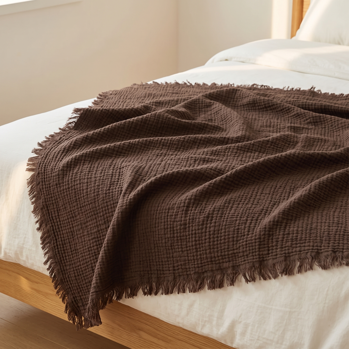 Crinkled Cotton Blanket with Fringe in Dark Brown – draped across the foot of a bed with white linen bedding