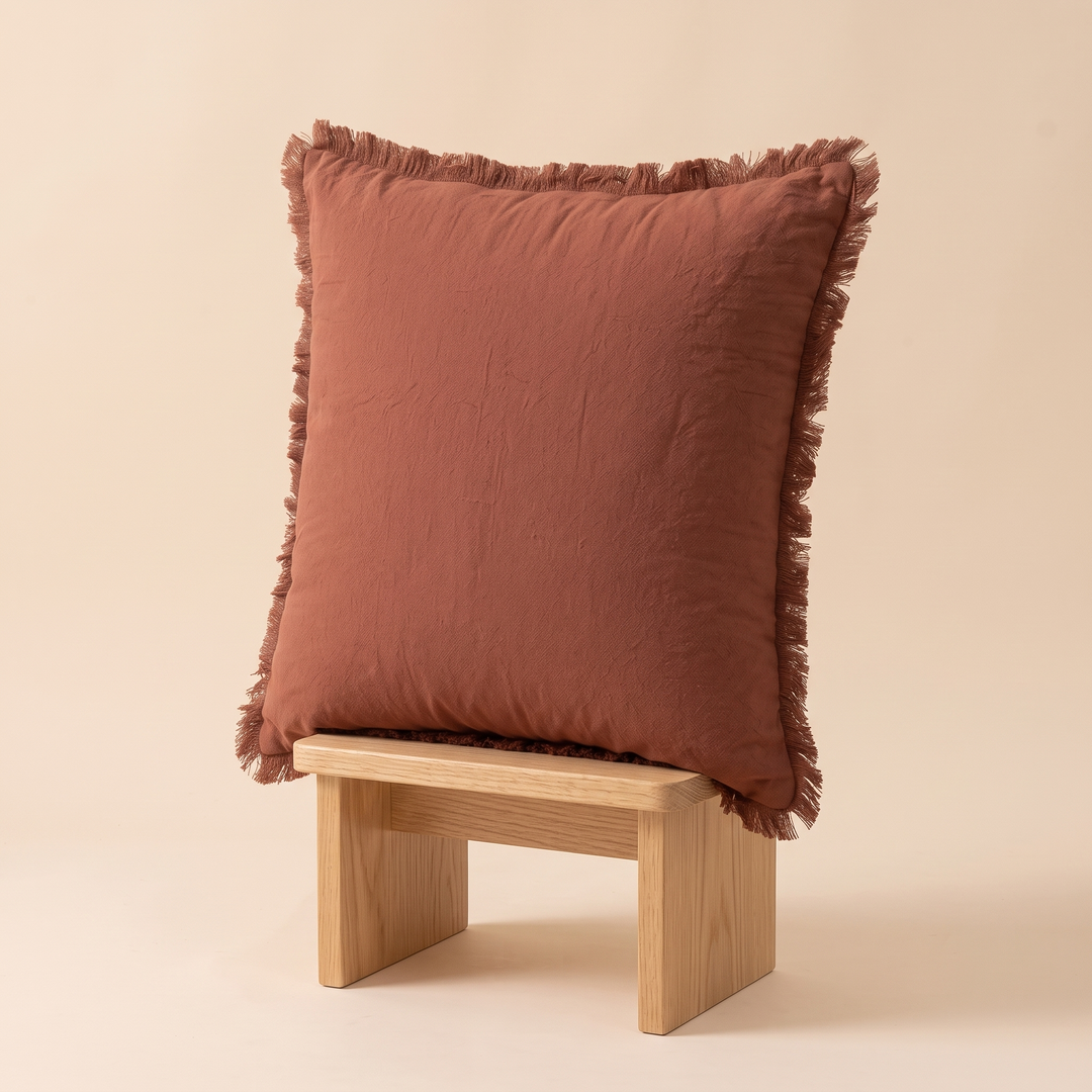 Fringed Pillow Cover in Chestnut Brown – propped upright on a small wooden stool against a peach backdrop