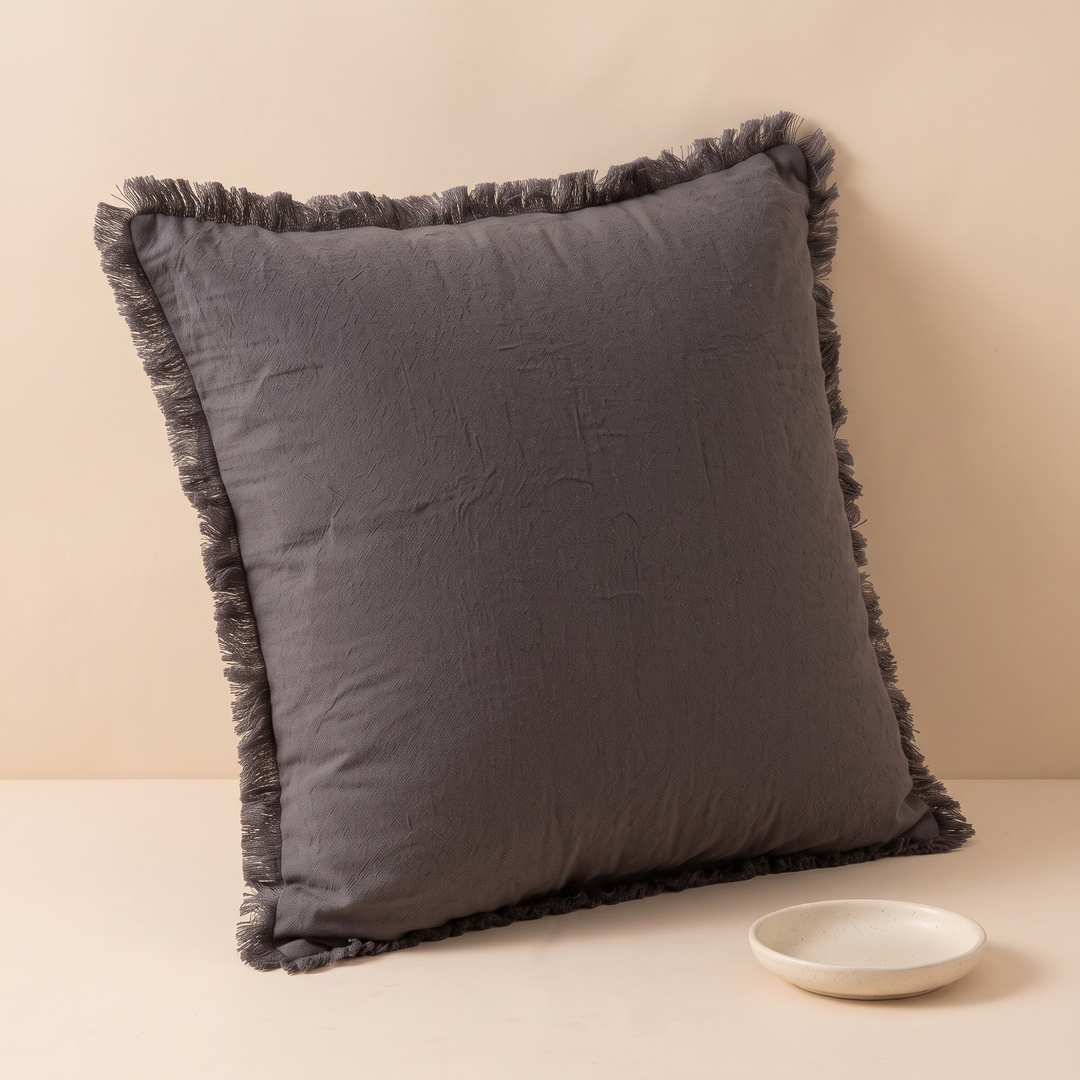 Fringed Pillow Cover in Dark Gray – leaning against a wall with a small speckled ceramic dish in front