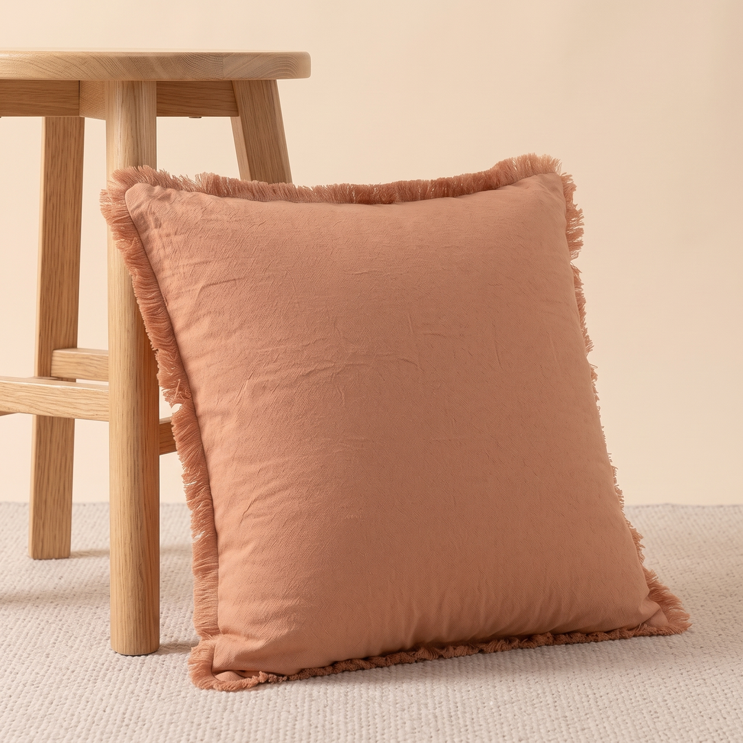 Fringed Pillow Cover in Orange – leaning against a wooden stool leg on a woven rug