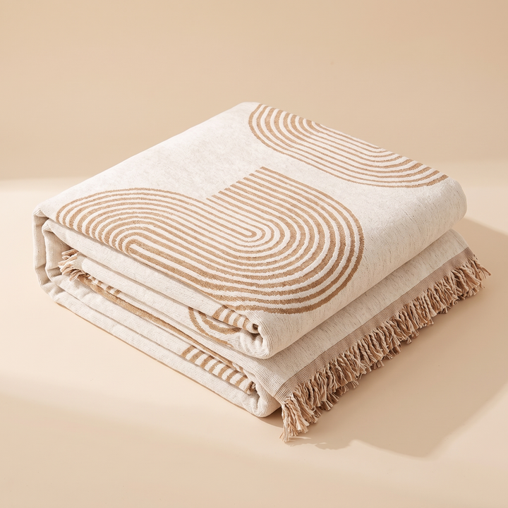 Geometric Chenille Couch Cover with Fringe in Beige – Arch – folded neatly in a stack on a plain peach backdrop