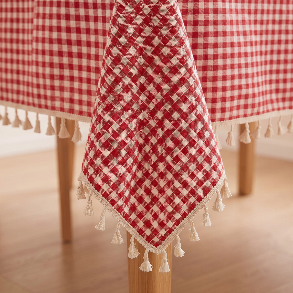 Red Gingham Tablecloth with Tassels in Red – Gingham – corner hanging over a wooden table leg, tassel trim visible