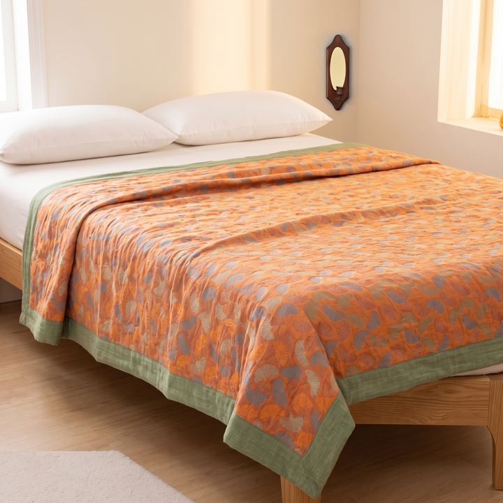 Ginkgo Leaf Gauze Blanket in Orange &amp; Green – draped over a wood-framed bed beside a sunlit window