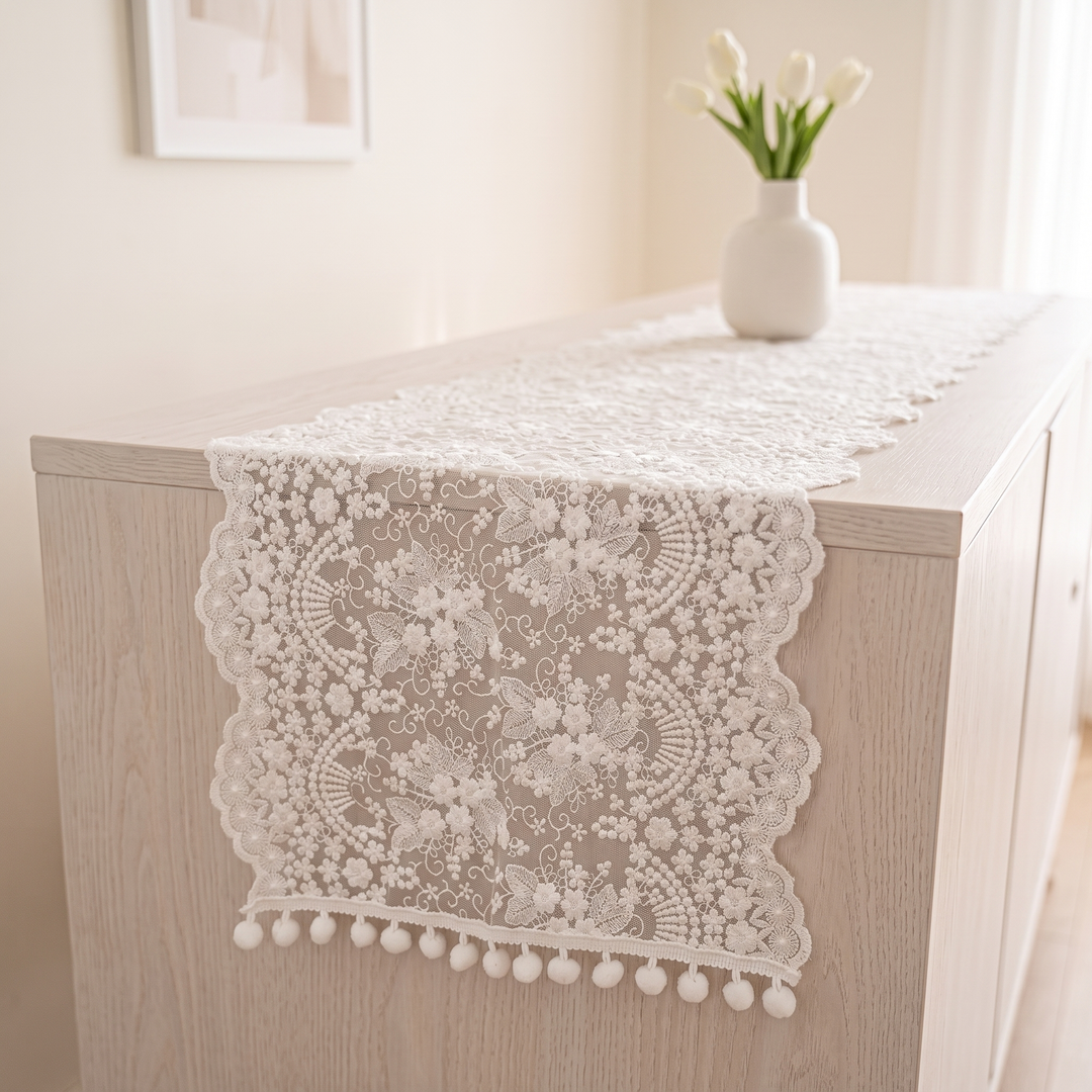 Lace Table Runner in White – Pom-Pom – hanging over the front edge of a light wood sideboard beside a tulip vase