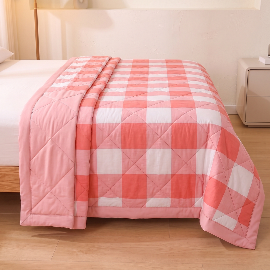 Lightweight Quilted Coverlet in Coral Buffalo Check – spread across a white-sheeted bed, side falling to wood floor