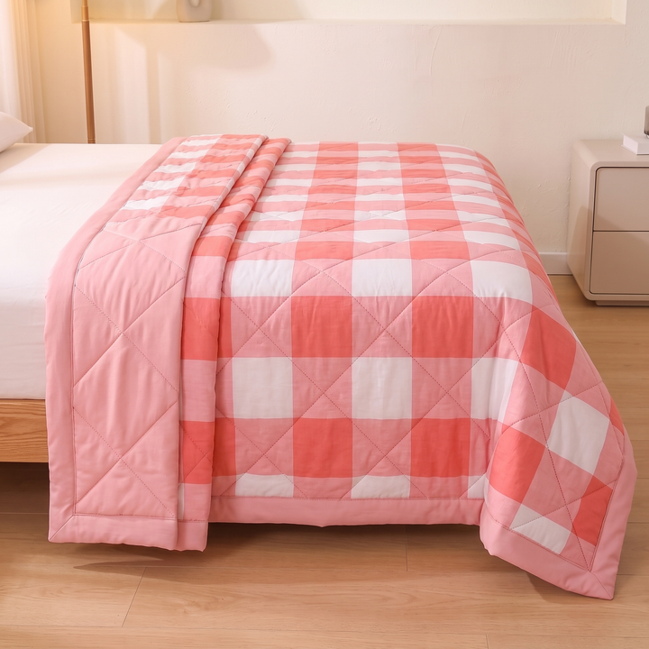 Lightweight Quilted Coverlet in Coral Buffalo Check – spread across a white-sheeted bed, side falling to wood floor