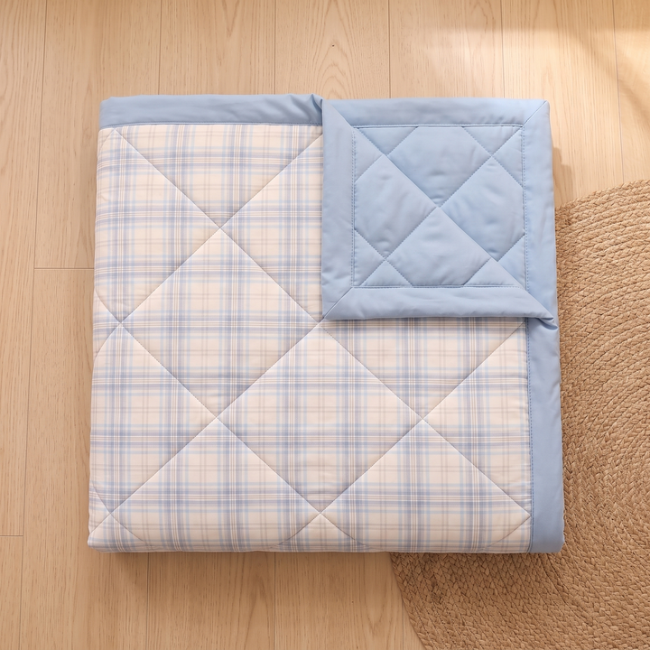 Lightweight Quilted Coverlet in Light Blue Plaid – top-down folded square on wood floor beside woven rug