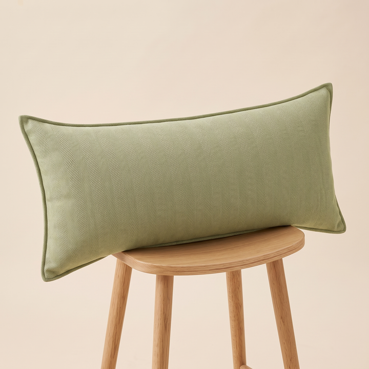 Long Lumbar Pillow Cover in Light Olive – resting sideways on a wooden stool against a peach wall