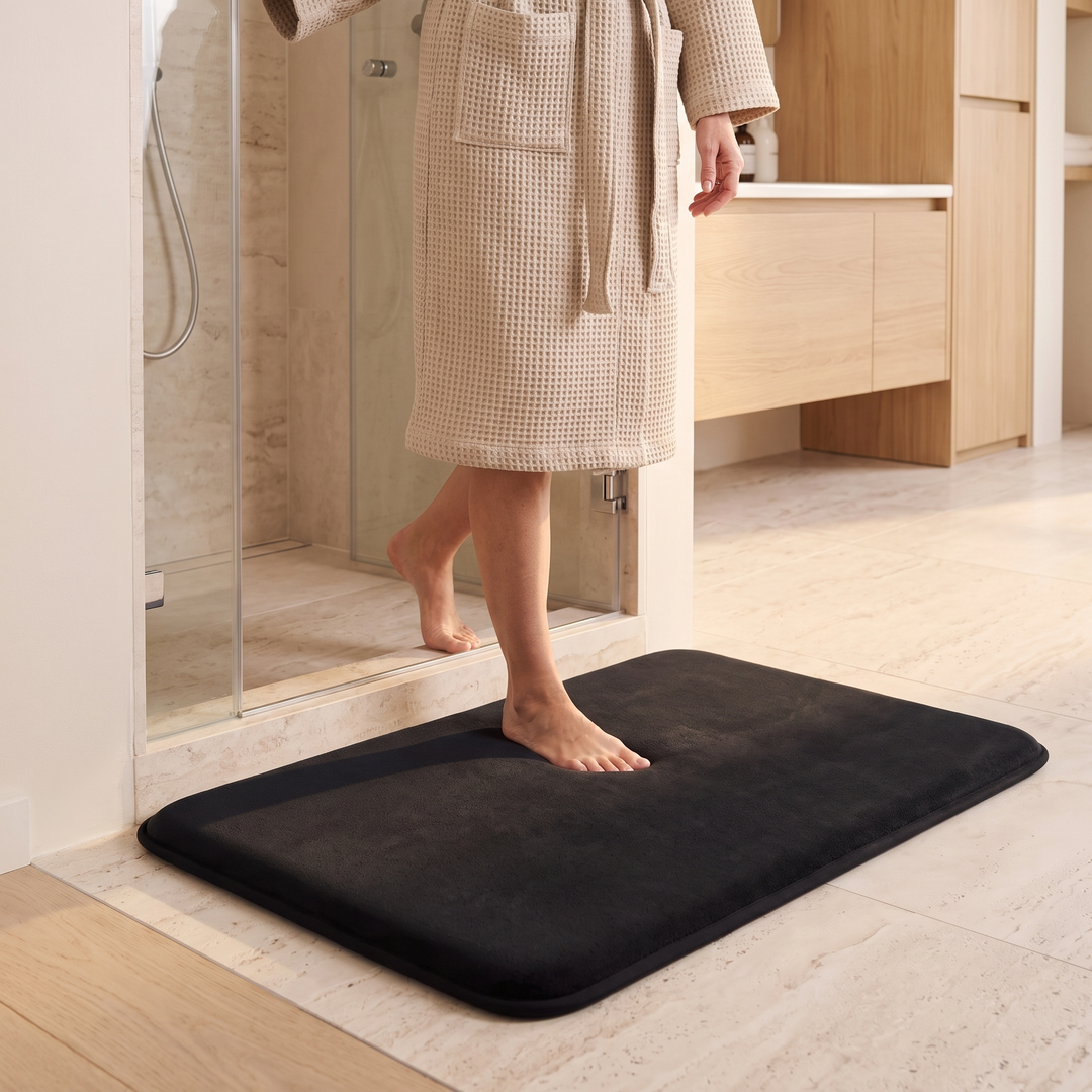 Non-Slip Memory Foam Bath Mat – Plush in Black – on a tiled floor outside a glass shower, person in robe stepping on