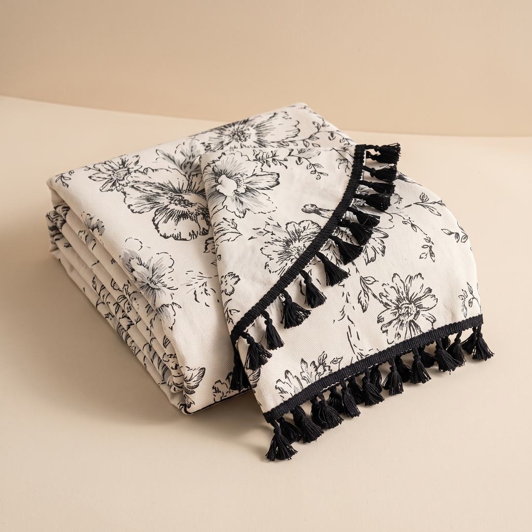 Round Floral Tablecloth in Black Floral – Tassels – folded in a neat stack on a plain beige surface