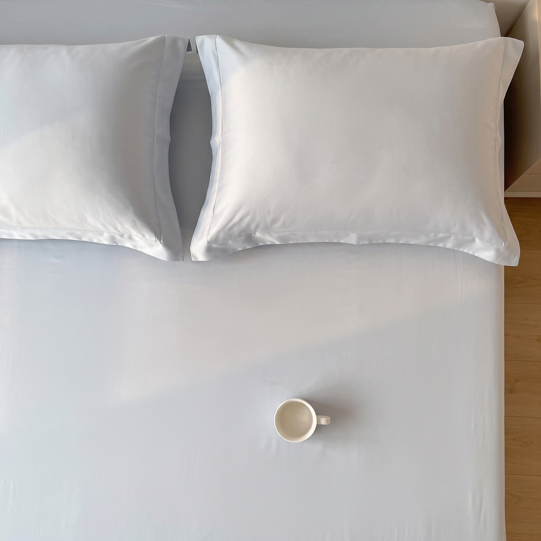 Sateen Fitted Sheet Set with 2 Pillowcases in Light Blue – top-down view of two pillows and a single teacup on the mattress
