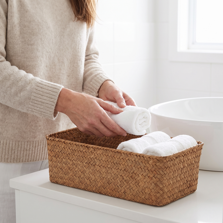 Seagrass Woven Basket – Bathroom Storage in Natural – on a white vanity as hands roll towels into it