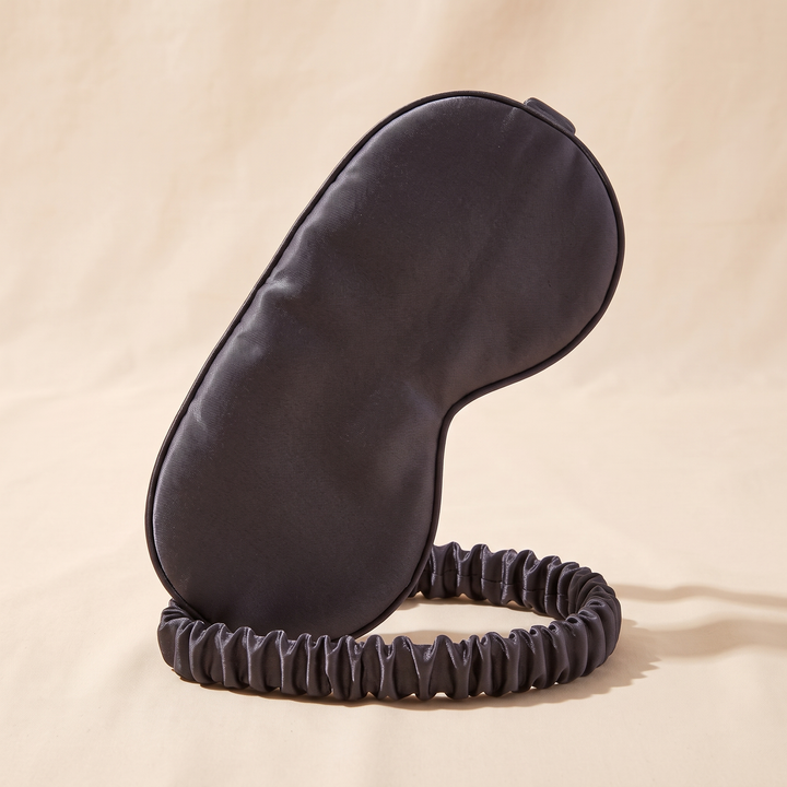Silk Sleep Mask in Dark Gray – standing upright on its elastic band against a peach backdrop