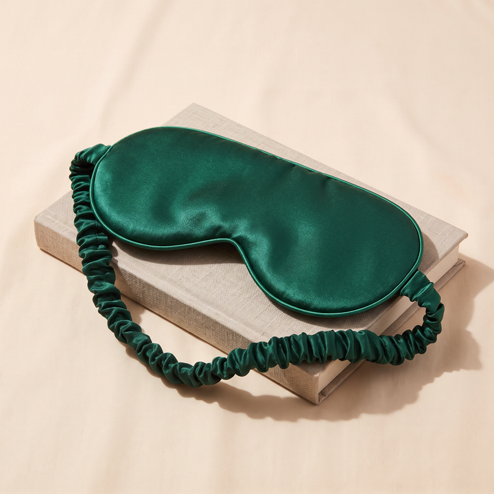 Silk Sleep Mask in Dark Green – resting on a closed linen-covered book, shot from above at an angle