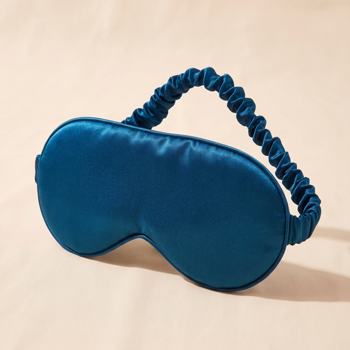 Silk Sleep Mask in Peacock Blue – lying with strap arched behind, crisp shadow to the right