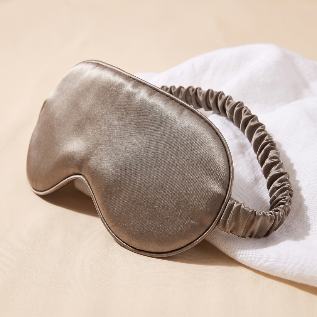 Silk Sleep Mask in Taupe – angled view resting on crumpled white linen against a beige backdrop