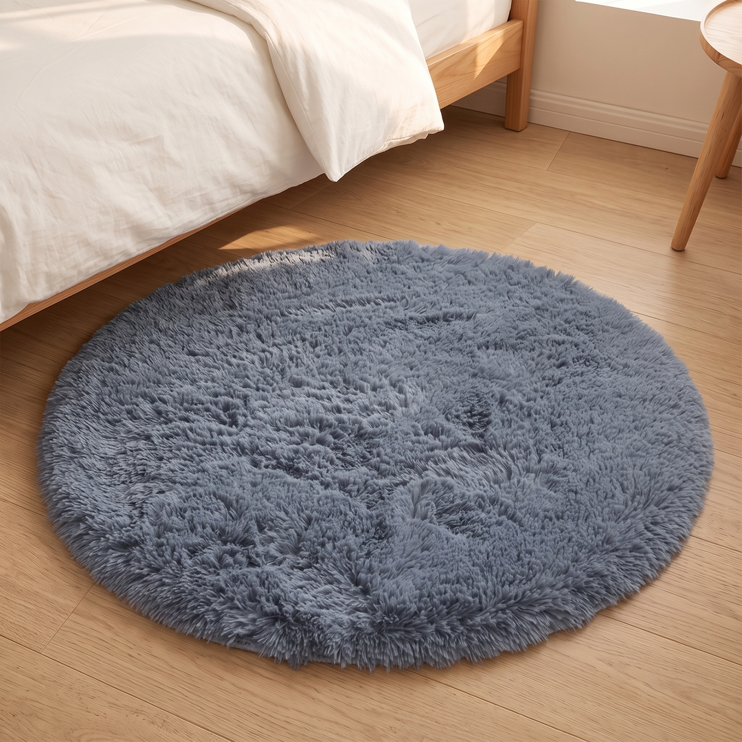 Small Round Shag Rug in Slate Blue – beside a white duvet and wooden bed frame in sunlight