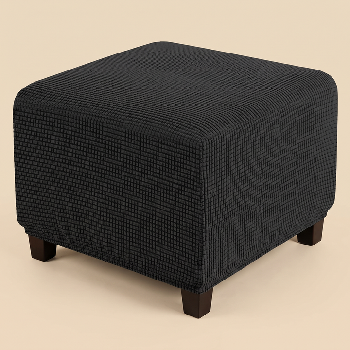 Stretch Ottoman Cover in Black – fitted on a square ottoman with dark wooden legs, plain beige background