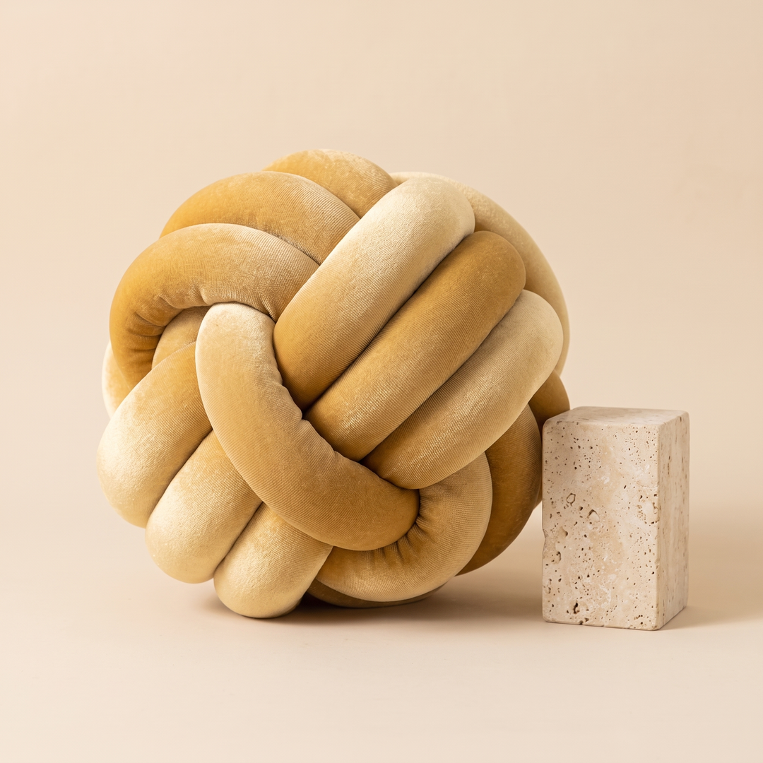 Velvet Knot Pillow in Honey Yellow – next to a travertine block against a plain peach backdrop