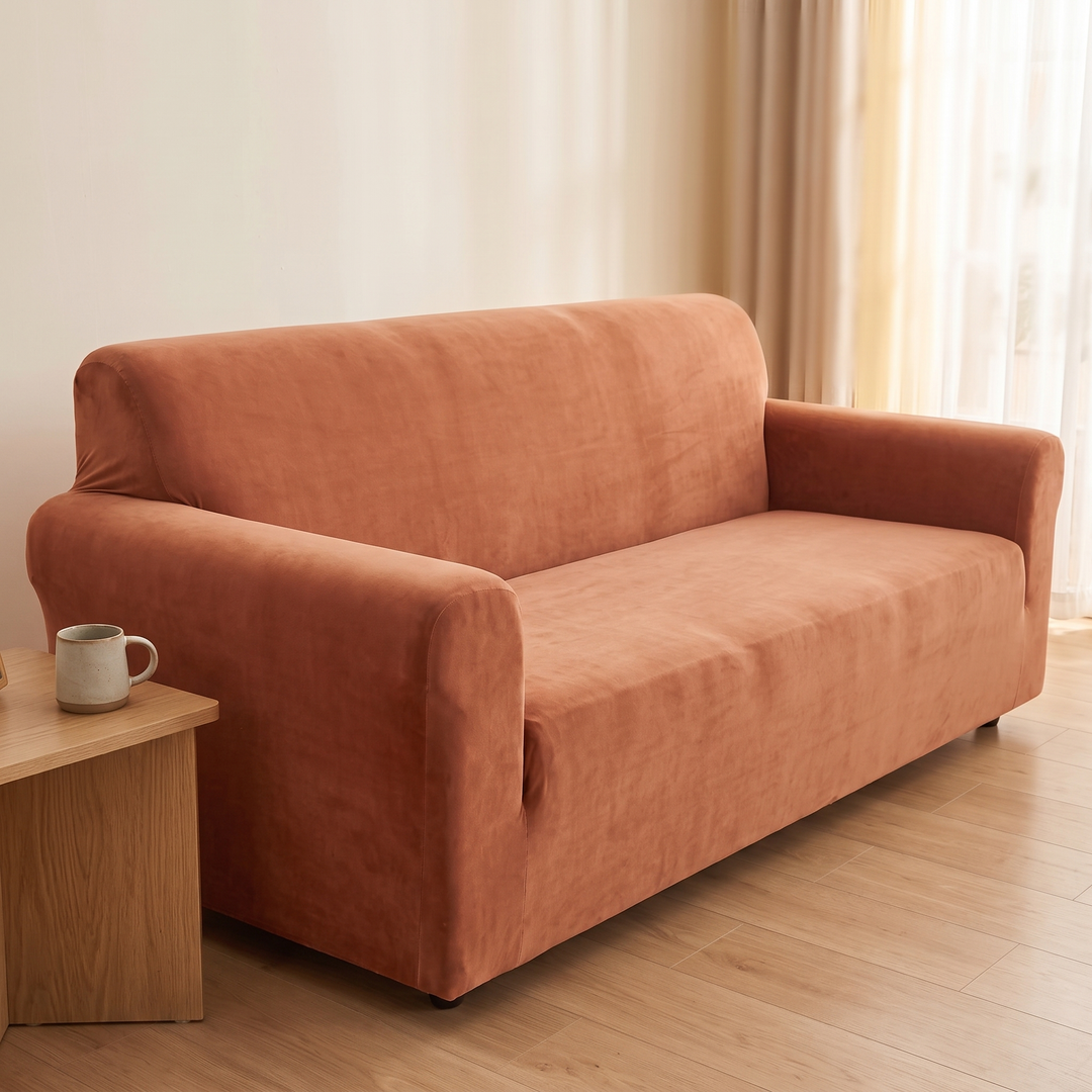 Velvet Stretch Sofa Cover in Rust – mug on a side table at left, curtains at right