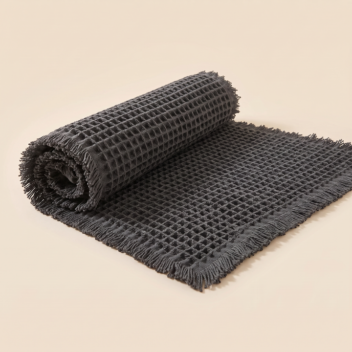 Waffle Bath Mat with Tassels in Dark Gray – partly rolled up from one end with fringe visible