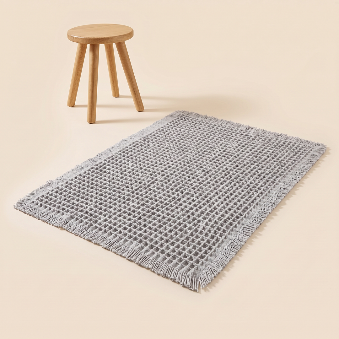 Waffle Bath Mat with Tassels in Light Gray – spread out next to a small round wooden stool
