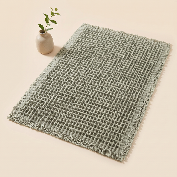 Waffle Bath Mat with Tassels in Sage – beside a small ceramic bud vase with a leafy stem