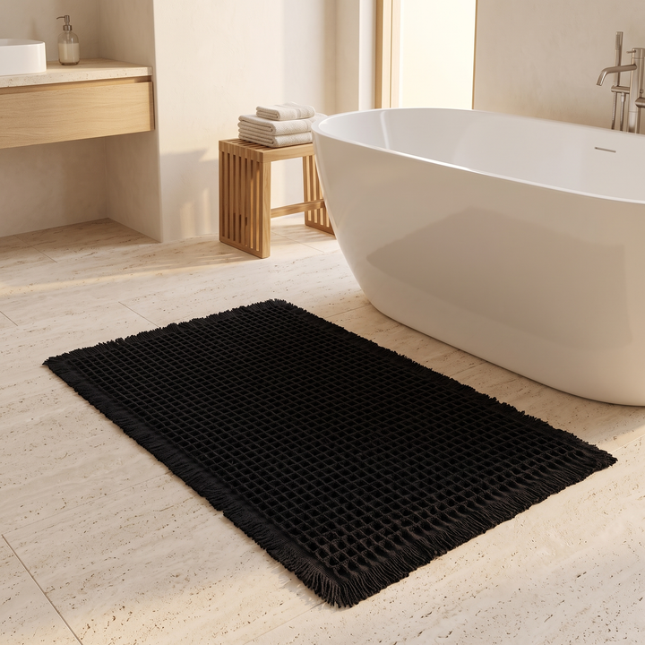 Waffle Bath Mat with Tassels in Black – on a travertine bathroom floor beside a freestanding tub