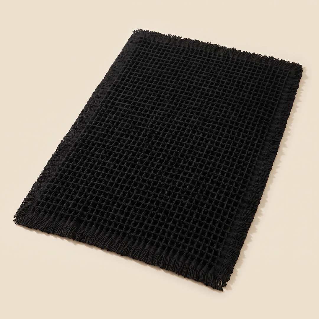 Waffle Bath Mat with Tassels in Black – laid flat on a plain cream background, seen from above at an angle