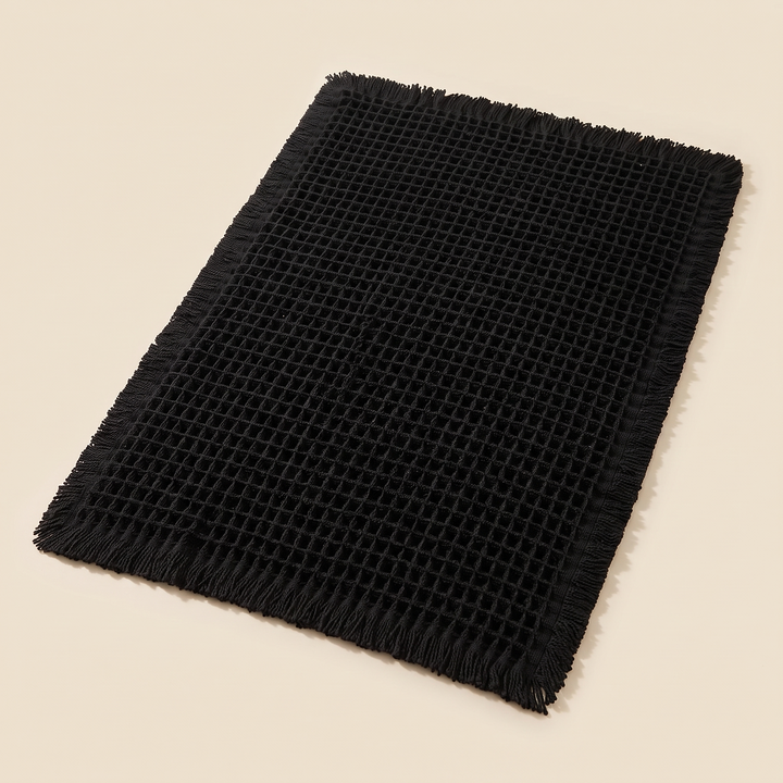 Waffle Bath Mat with Tassels in Black – laid flat on a plain cream background, seen from above at an angle