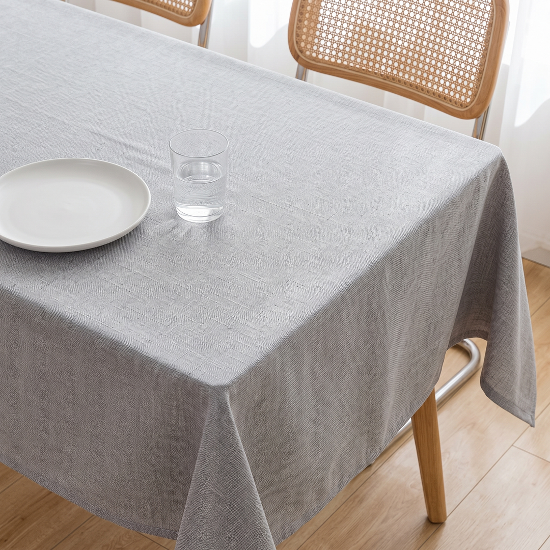 Wipeable Tablecloth – Linen Look in Gray – overhanging a table beside cane-back chairs near sheer curtains