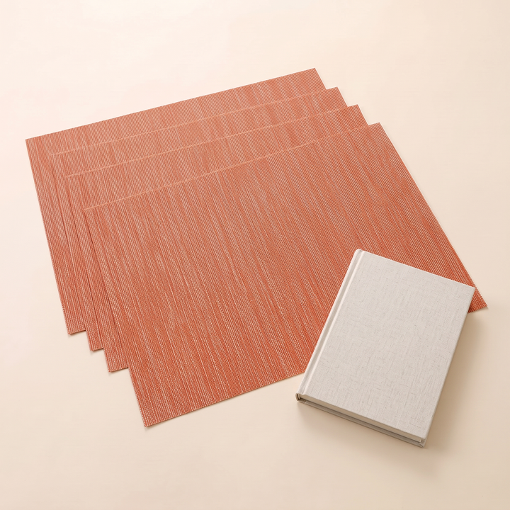 Woven Placemats – Wipe Clean in Orange – Crosshatch – overlapped with a closed linen-covered book on one corner